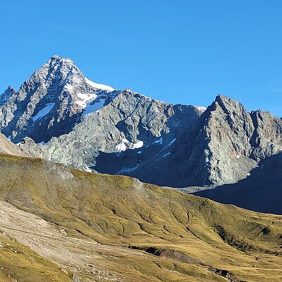 Berglandschaft mit schneebedecktem Gipfel, felsigen Hängen und grünem Tal unter klarem, blauem Himmel. Der Berg ist der Großglockner
